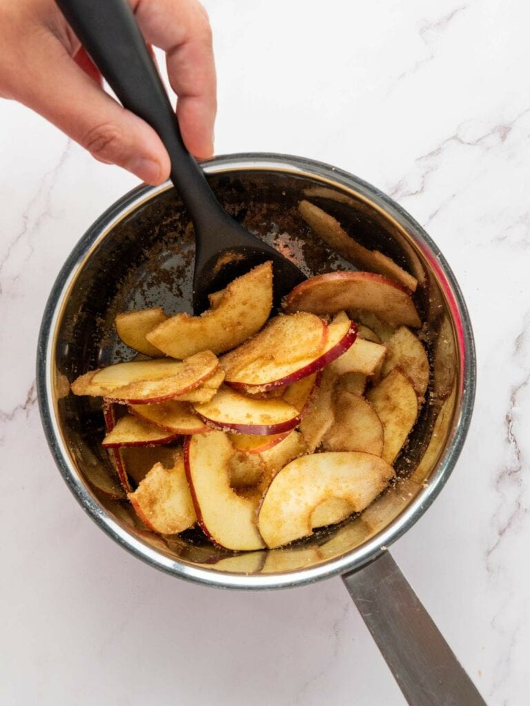 A hand uses a black spatula to mix sliced apples coated with cinnamon and sugar in a stainless steel saucepan on a marble surface.