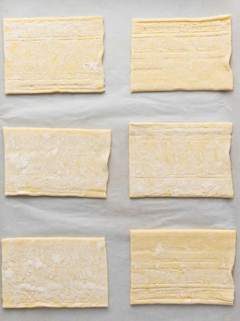 Six rectangular pieces of uncooked puff pastry dough are evenly spaced on a sheet of parchment paper.
