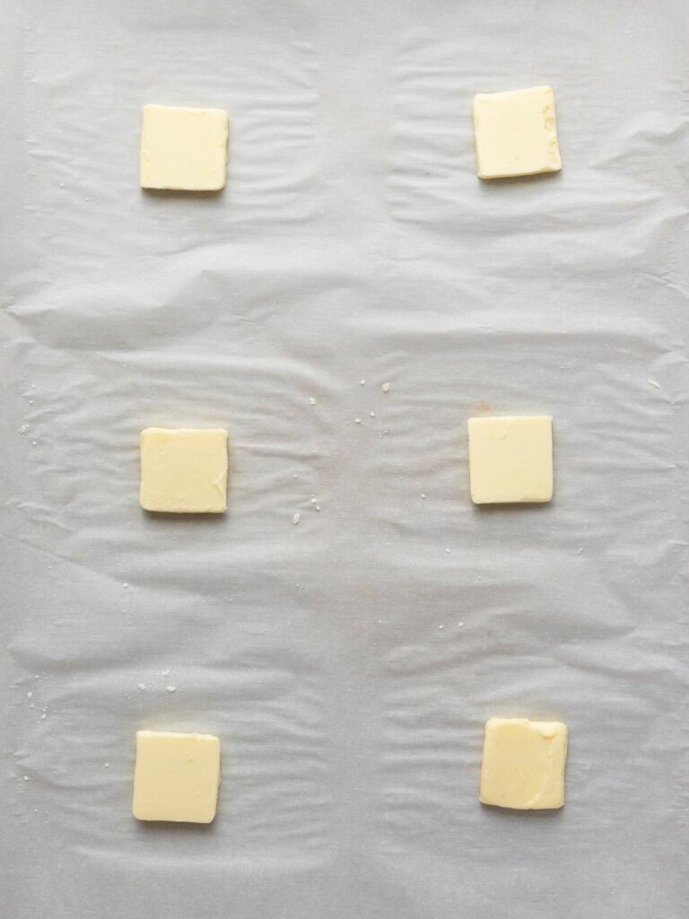 Six square slices of butter evenly spaced on a sheet of parchment paper.