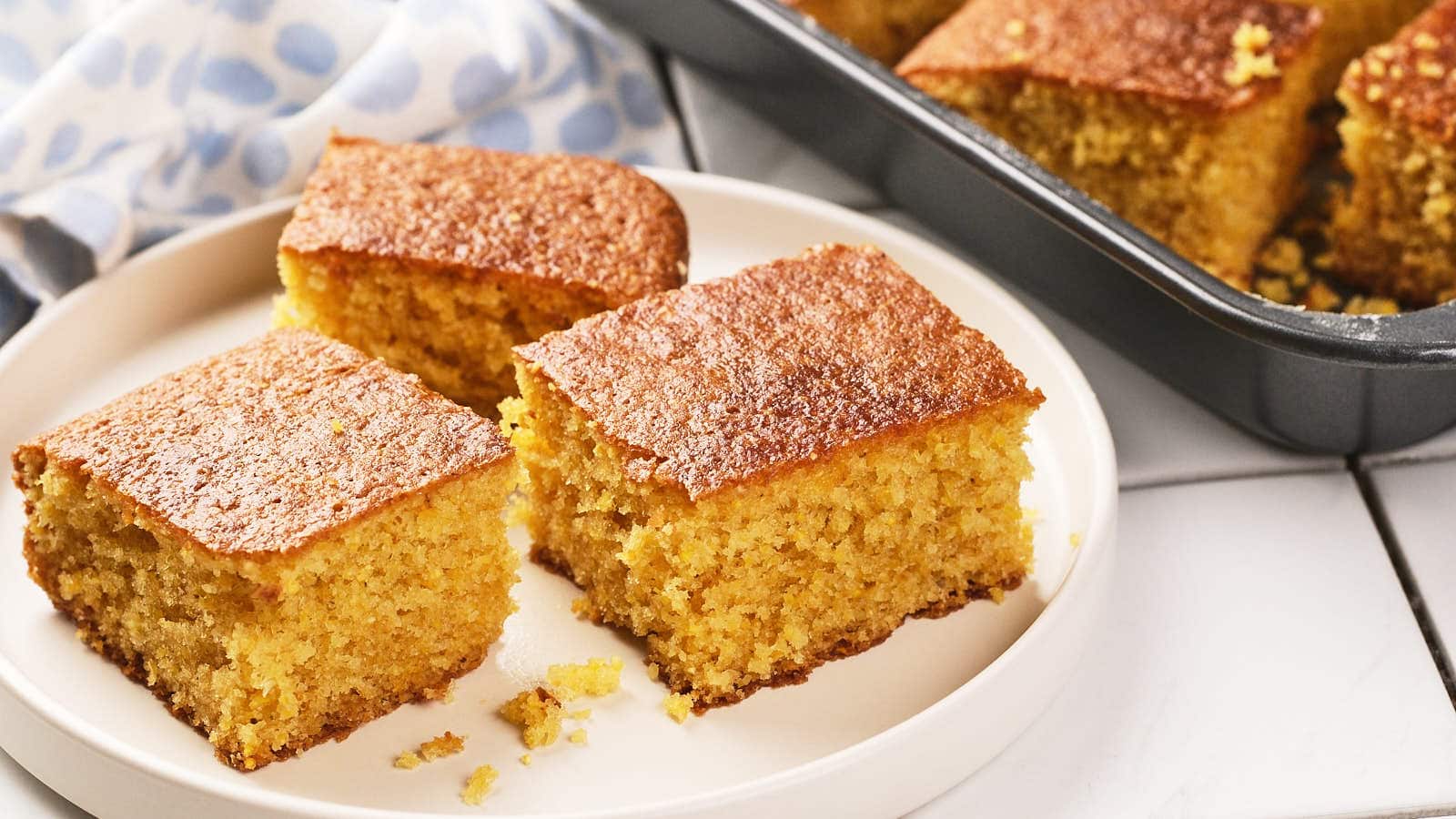 Three square pieces of cornbread are on a white plate, with more pieces visible in a baking pan in the background.