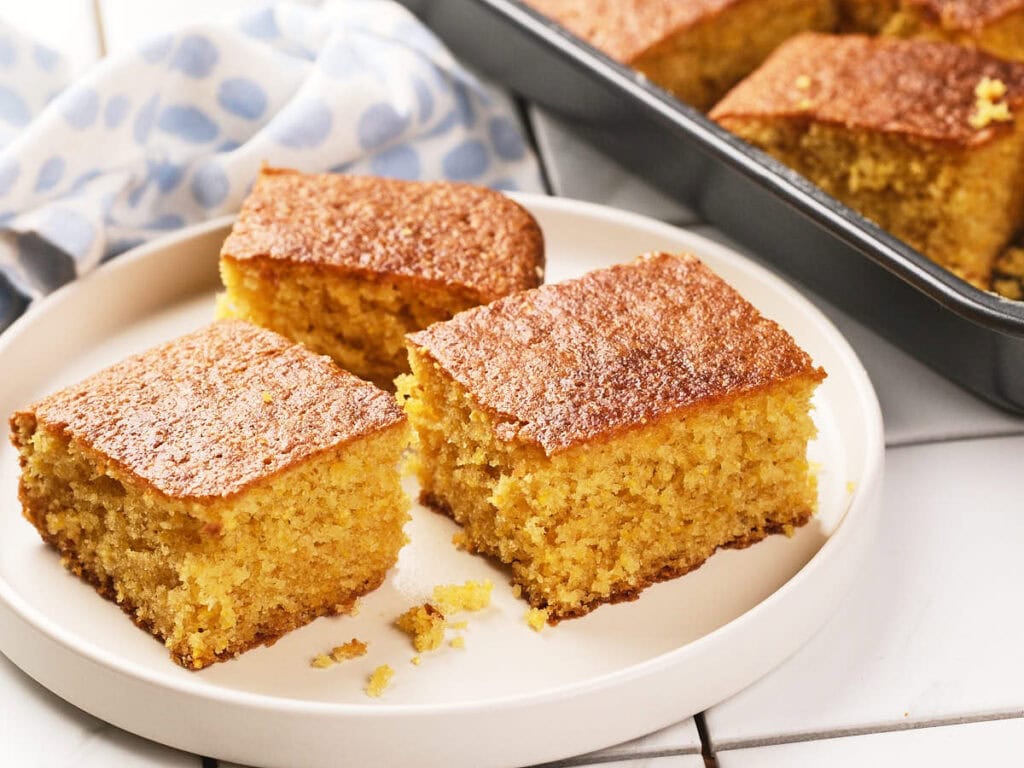 Three pieces of cornbread are served on a white plate, with a baking tray of more cornbread and a blue and white cloth in the background.