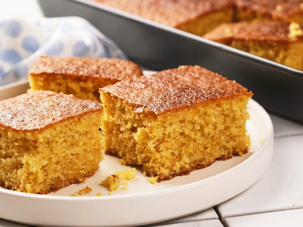 Three pieces of cornbread are served on a white plate, with a baking pan containing more cornbread in the background.
