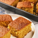 Three pieces of cornbread are served on a white plate in front of a baking pan filled with more cornbread.