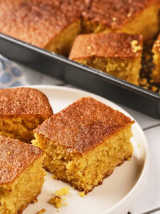Three pieces of cornbread are served on a white plate in front of a baking pan filled with more cornbread.