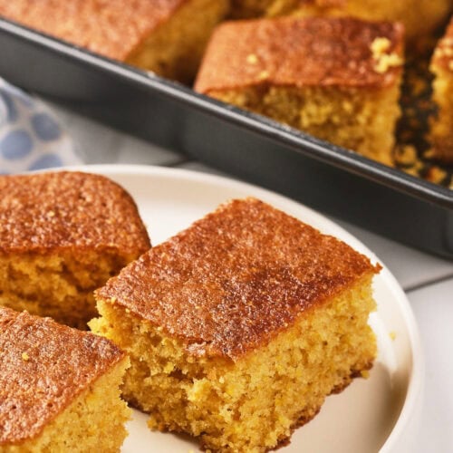 Three pieces of cornbread are served on a white plate in front of a baking pan filled with more cornbread.