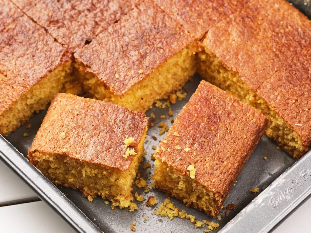 A baking pan filled with rectangular slices of golden-brown cornbread, some pieces removed and crumbs visible in the pan.