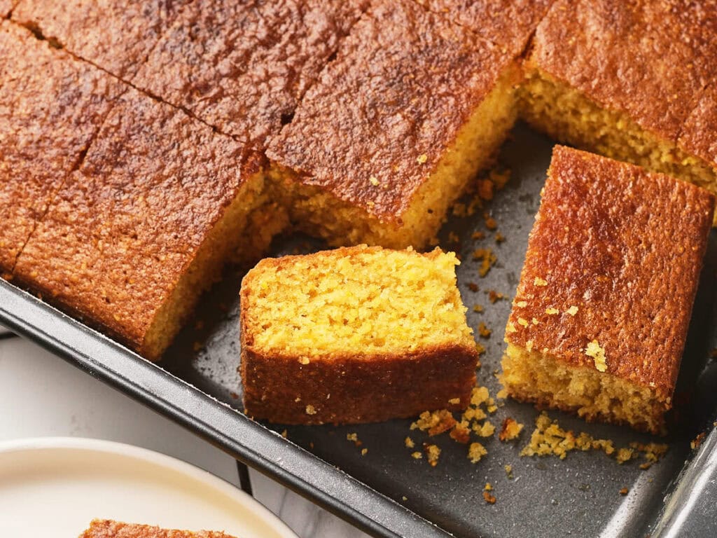 A pan of golden brown cornbread cut into squares, with one piece partially removed and crumbs scattered on the tray.