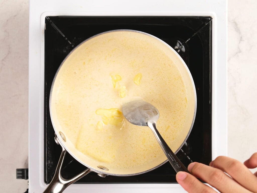 A hand stirring melted butter into a creamy mixture in a saucepan on a stovetop.
