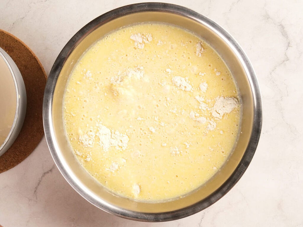 A stainless steel bowl containing a yellow batter mixture with visible lumps of flour on its surface.
