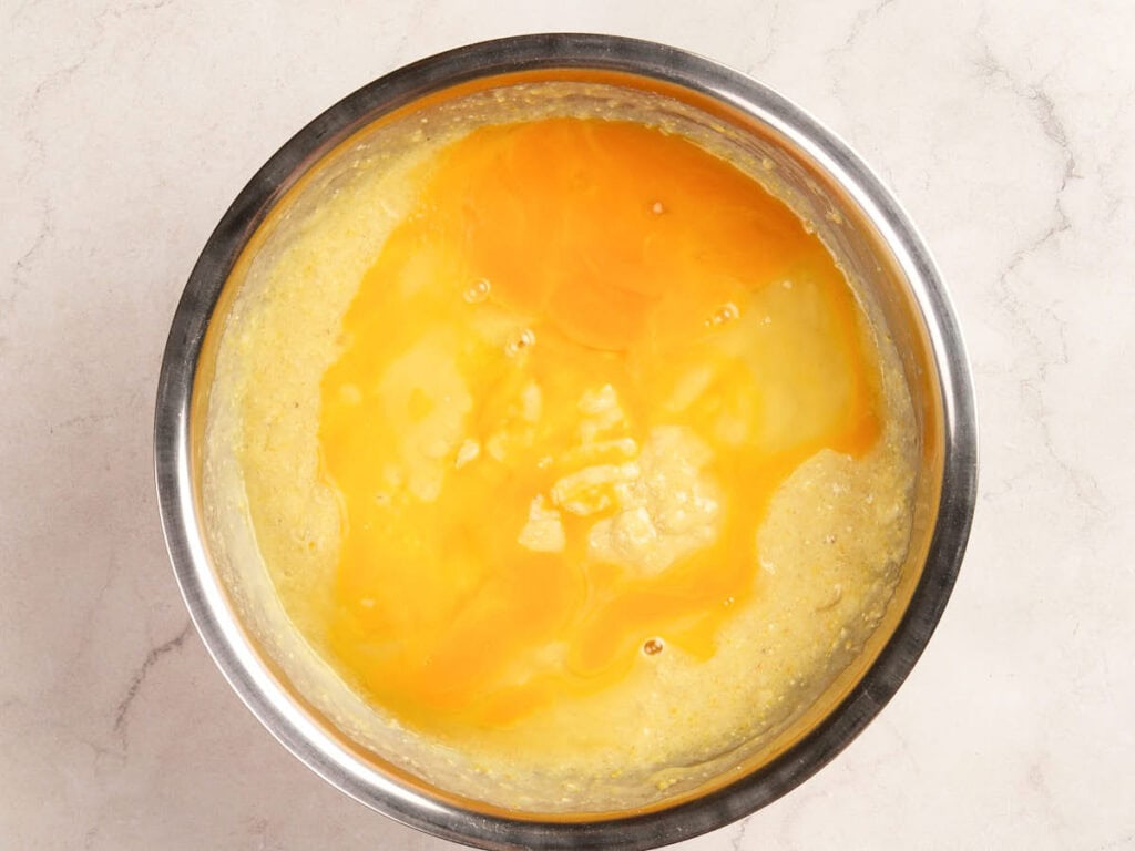 A stainless steel bowl containing a yellow batter mixture with cracked raw eggs on top.
