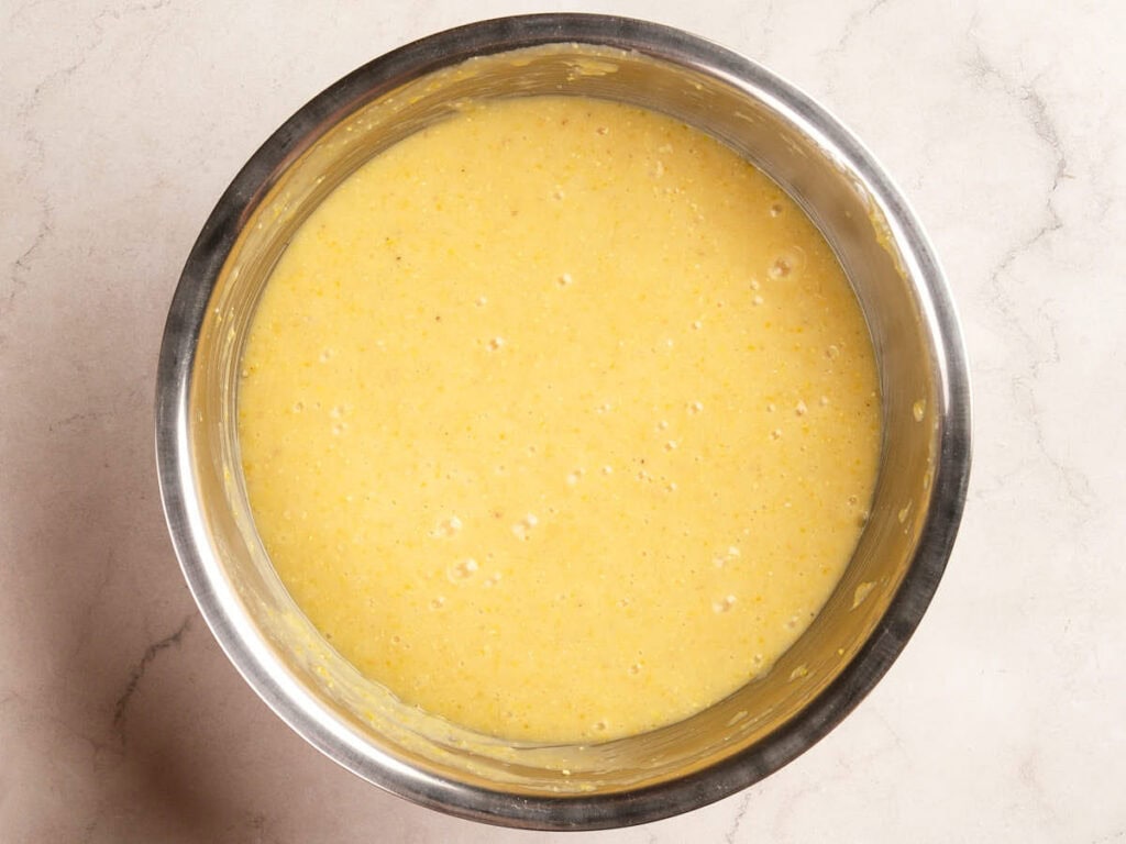 A stainless steel bowl filled with yellow cornbread batter sits on a light marble countertop.