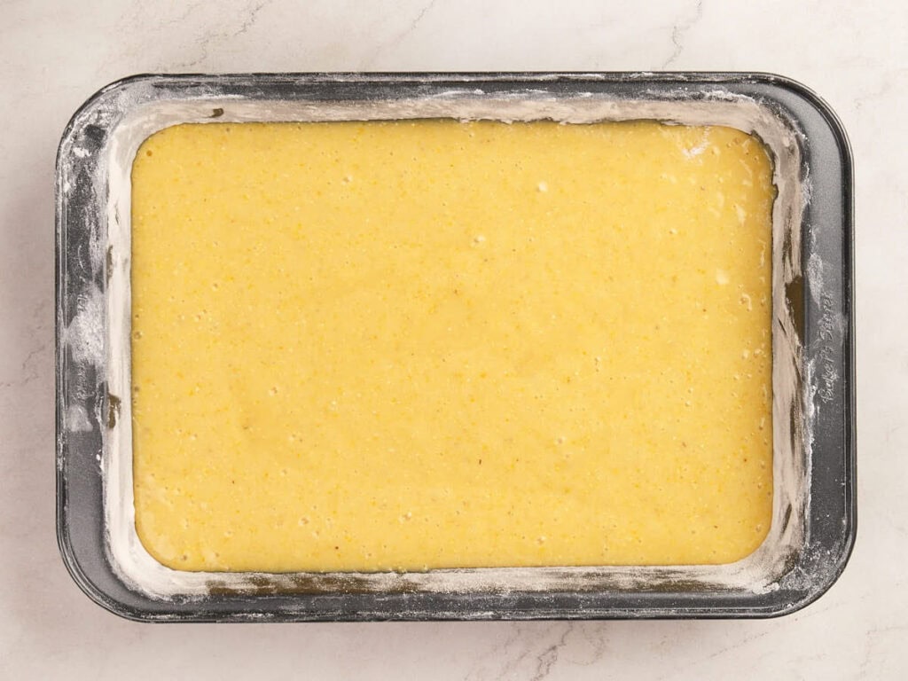 Rectangular metal baking pan filled with an unbaked yellow cake batter, with edges lightly dusted in flour, on a white countertop.
