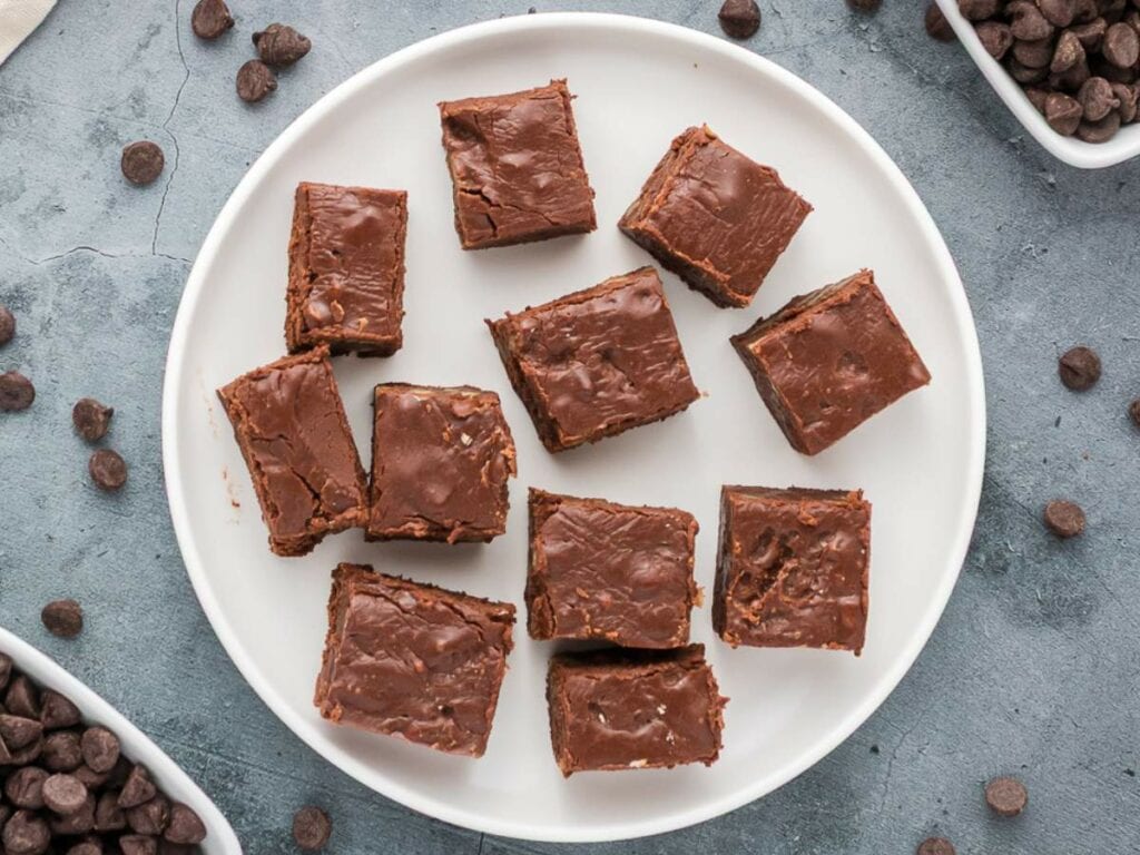 A white plate with twelve square pieces of chocolate fudge is on a gray surface, with chocolate chips scattered around.