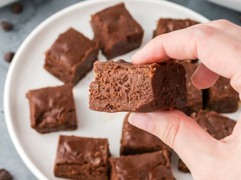 A hand holds a partially eaten chocolate brownie above a white plate with several whole brownies.