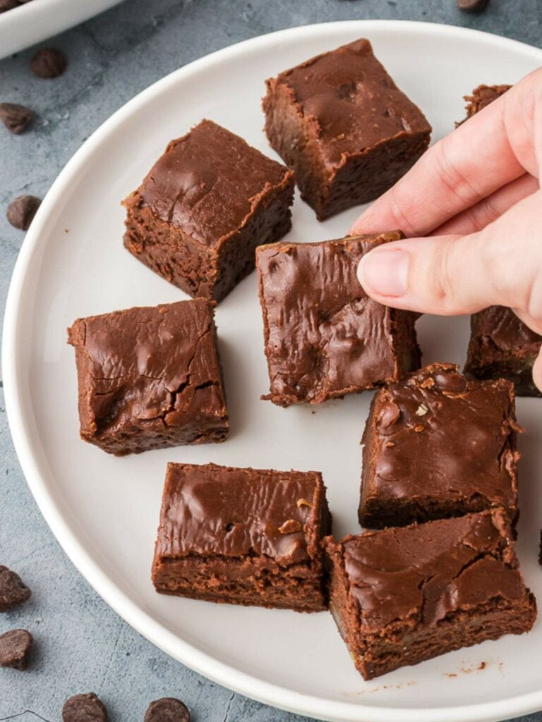 A hand reaching to pick up a square piece of chocolate fudge from a white plate with several other fudge pieces.