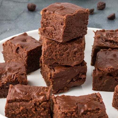 A white plate with several pieces of chocolate fudge stacked and arranged, with a few chocolate chips scattered in the background.