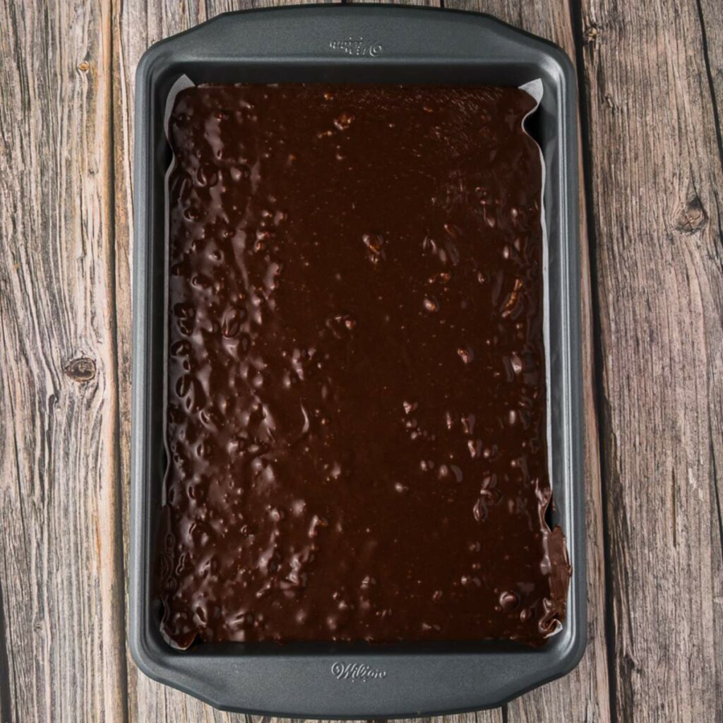 A baking pan filled with unbaked chocolate brownie batter, resting on a wooden surface.