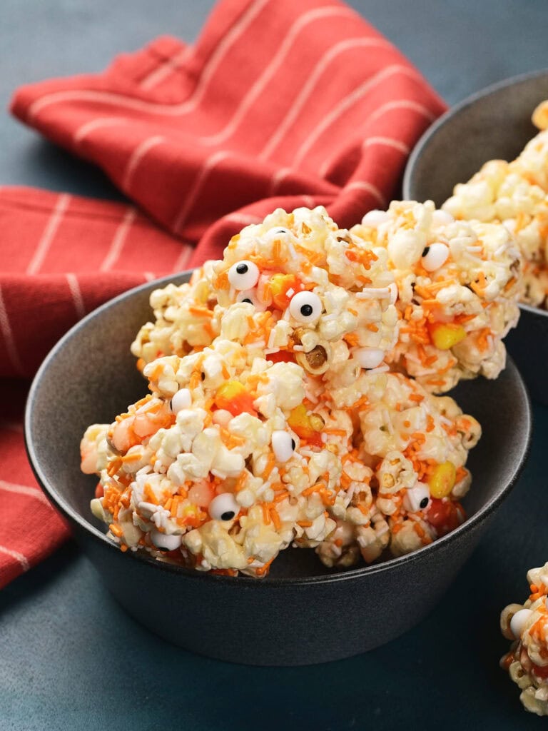 A bowl of popcorn clusters mixed with candy corn and candy eyes, placed in front of a red striped cloth.