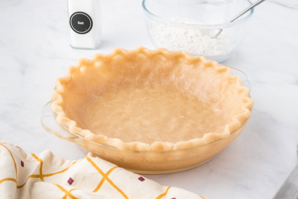 A glass pie dish with an unbaked pie crust, next to a bowl of flour with a spoon, a container of salt, and a patterned kitchen towel on a white surface.