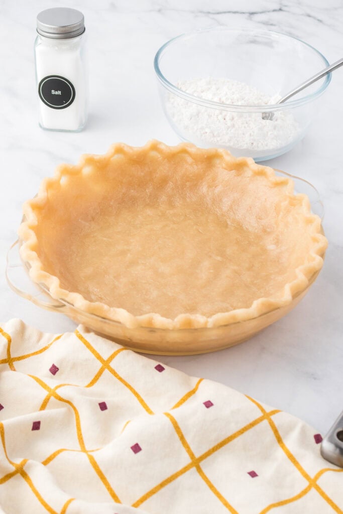 An unbaked pie crust in a glass pie dish sits on a counter next to a bowl of flour with a spoon, a salt container, and a patterned kitchen towel.