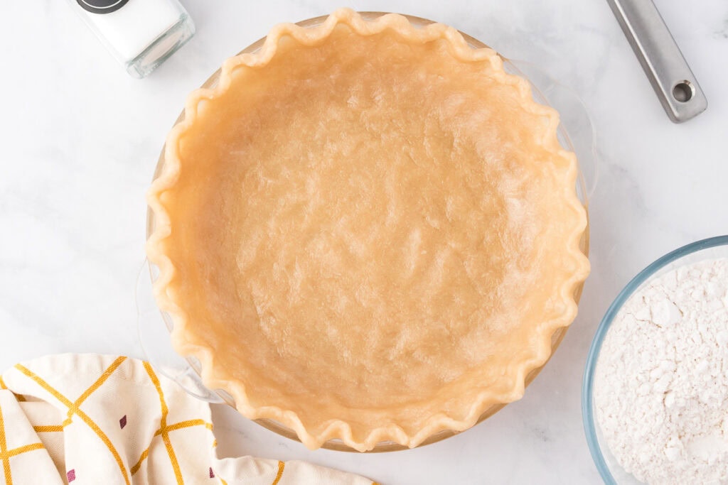 Unbaked pie crust in a glass pie dish, surrounded by a bowl of flour, a salt shaker, a metal utensil, and a cloth on a marble countertop.