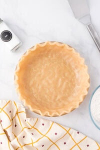 Unbaked pie crust in a glass pie dish on a marble countertop, with a salt shaker, a bowl of flour, a spatula, and a patterned dish towel nearby.