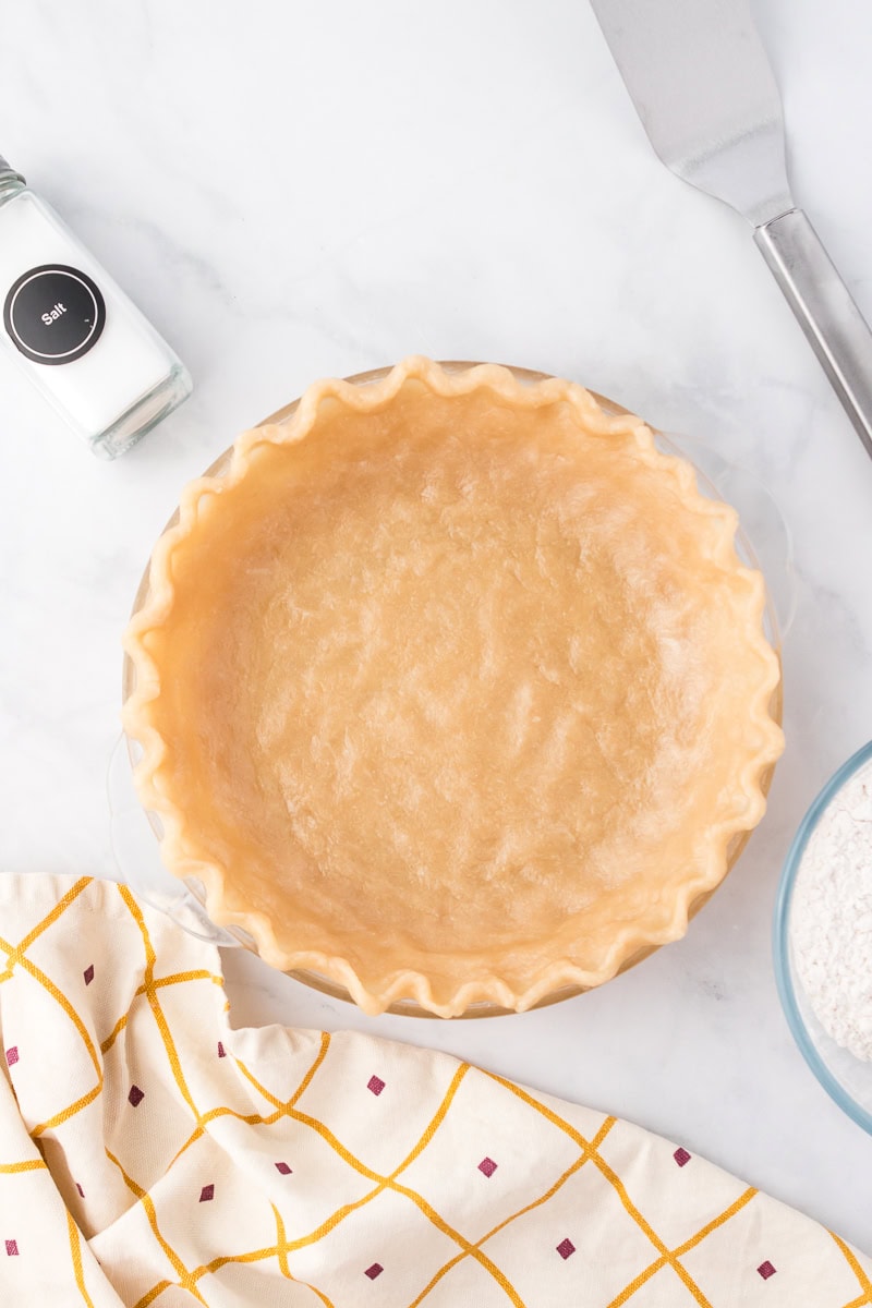 Unbaked pie crust in a glass pie dish on a marble countertop, with a salt shaker, a bowl of flour, a spatula, and a patterned dish towel nearby.