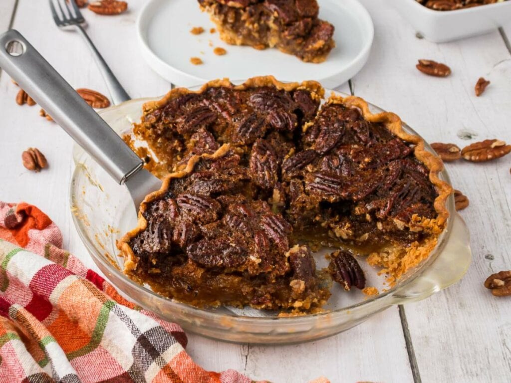 A partially sliced pecan pie in a glass dish with a serving spatula, surrounded by pecans and a colorful cloth on a wooden table.
