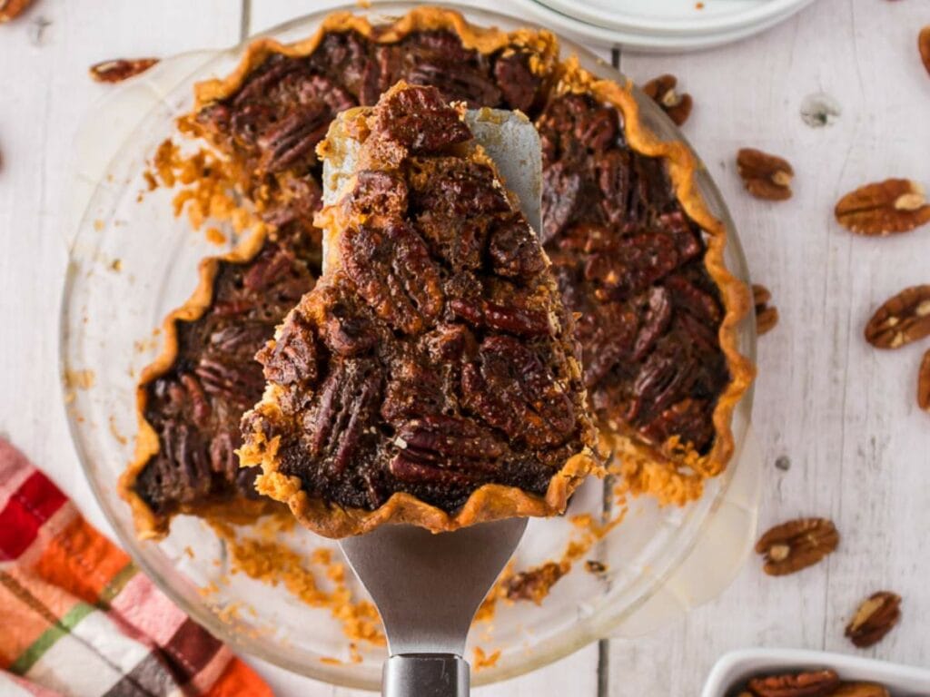 A slice of pecan pie is being lifted from a glass pie dish with a metal spatula on a wooden surface.