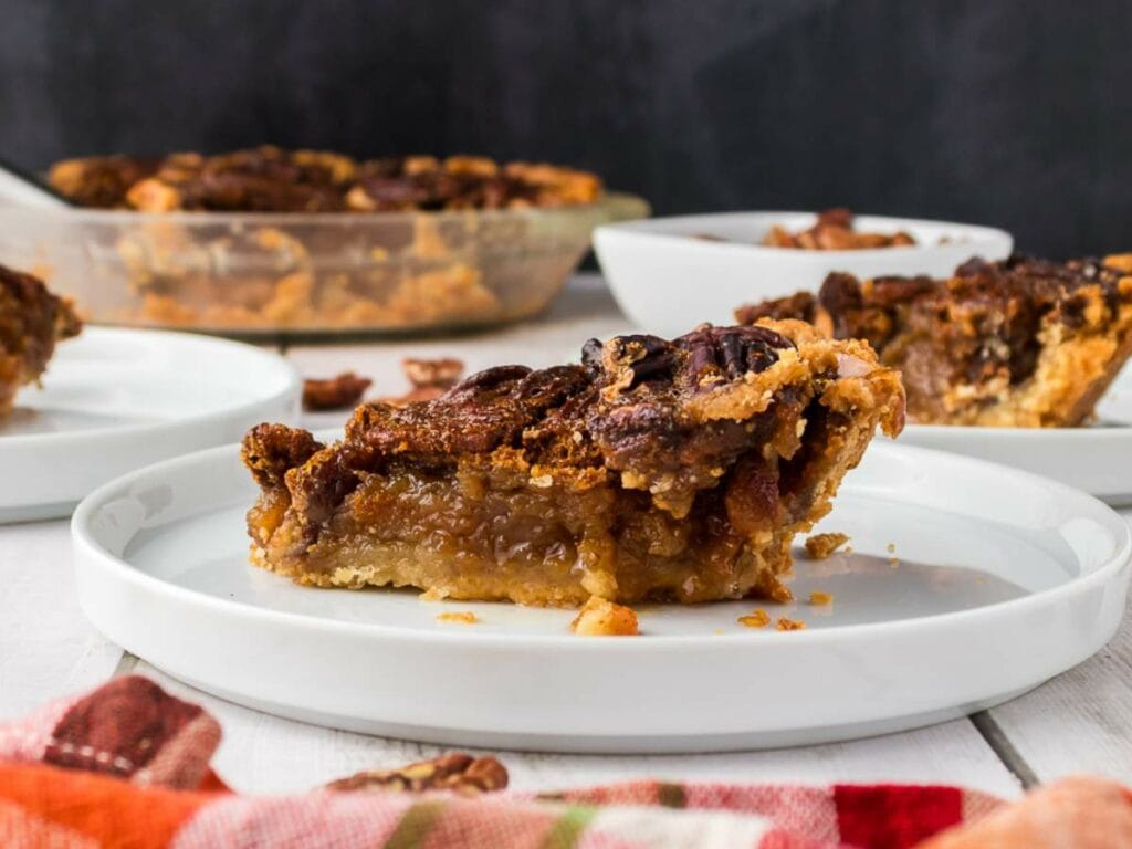 A slice of pecan pie sits on a white plate, with more pie and bowls in the background on a white surface.