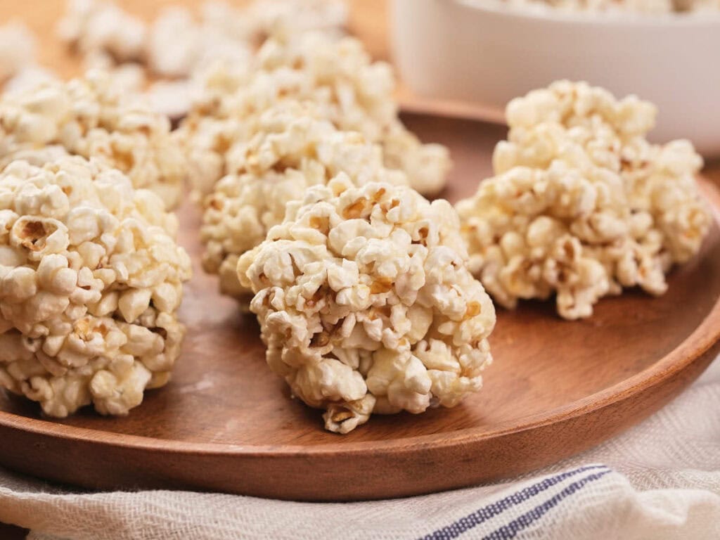 Several popcorn balls are arranged on a wooden plate, with a bowl partially visible in the background.