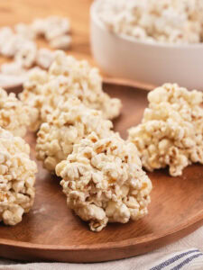 Several popcorn balls are arranged on a wooden plate, with a white bowl containing more popcorn in the background.