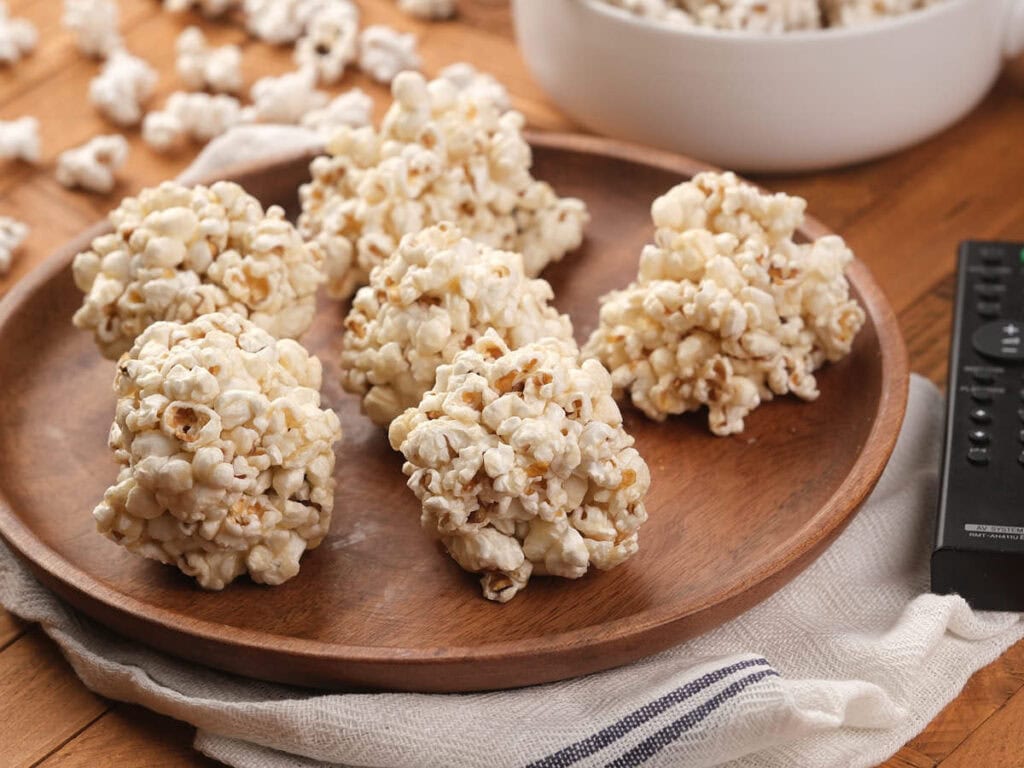 Five popcorn balls are arranged on a wooden plate atop a cloth napkin, with a bowl of more popcorn balls and a remote control visible nearby.