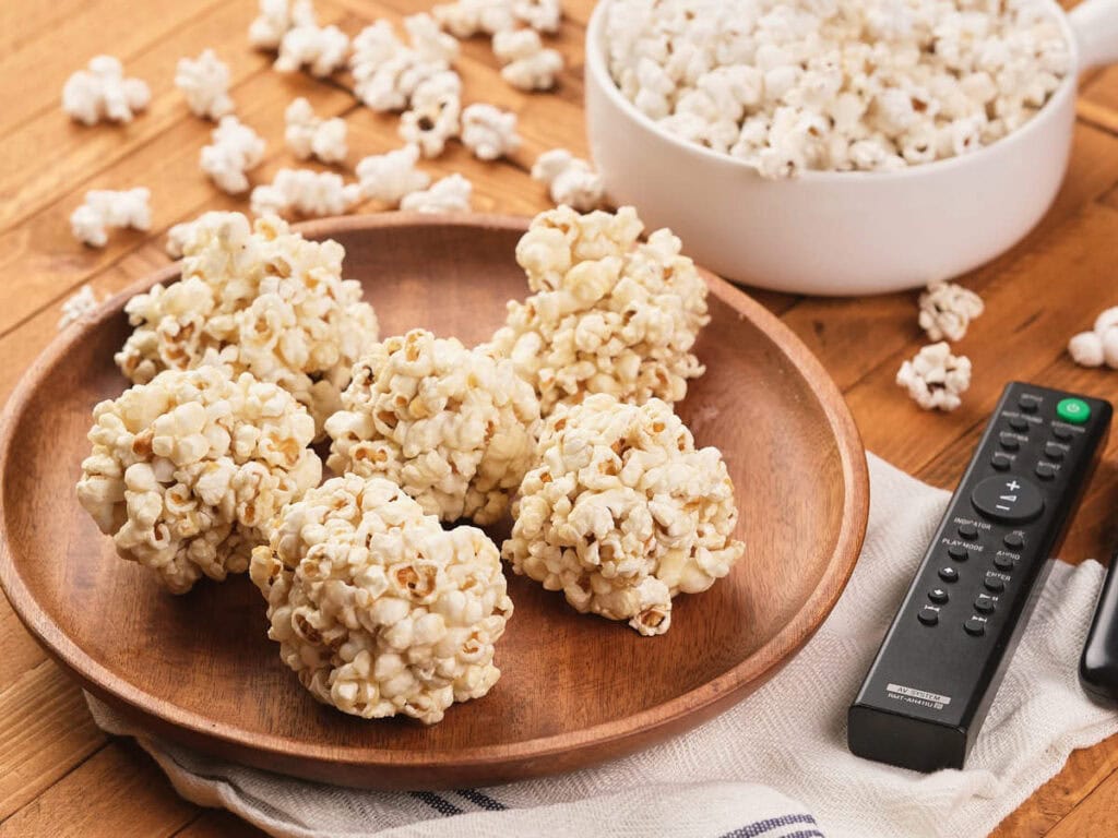 A wooden plate with five popcorn balls sits next to a bowl of popcorn and TV remotes on a wooden surface.