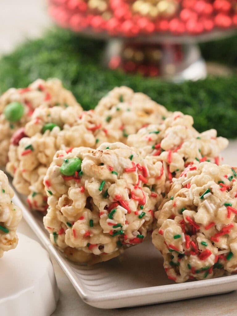 Several popcorn balls with red, green, and white holiday sprinkles and candy pieces are arranged on a white plate with festive decorations in the background.