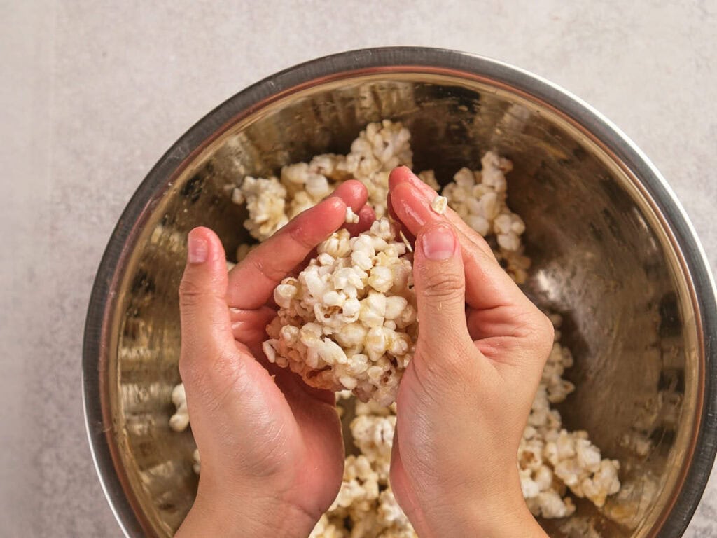 Hands shaping popcorn into a ball over a metal mixing bowl filled with popcorn on a light countertop.