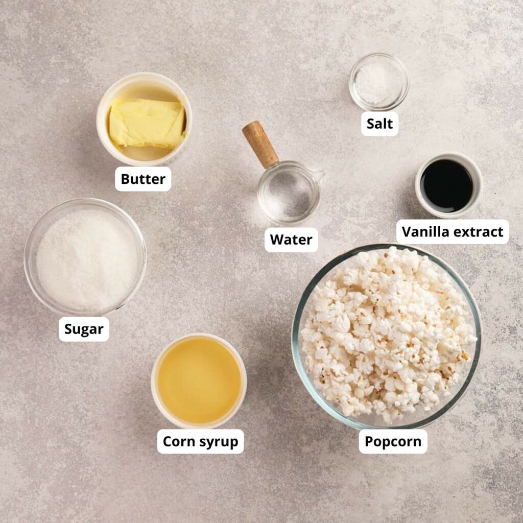 Seven labeled ingredients for making popcorn balls are arranged on a countertop: popcorn, sugar, butter, corn syrup, vanilla extract, salt, and water.