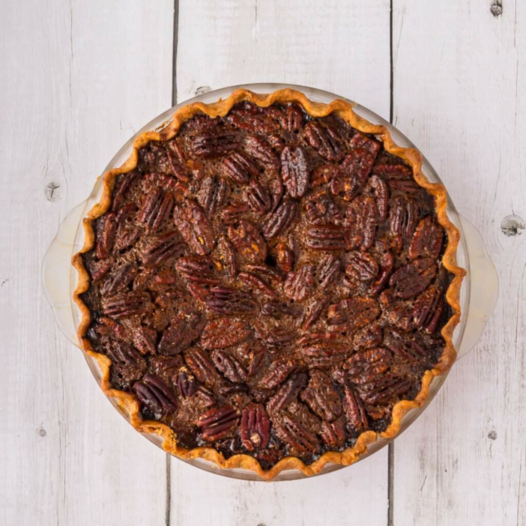 A whole pecan pie with a golden crust shown from above, placed on a white wooden surface.
