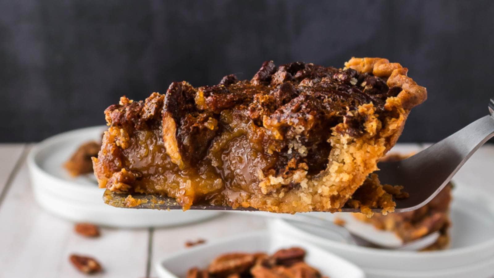 A close-up of a slice of pecan pie on a spatula, with a golden-brown crust and a gooey filling, held above a stack of white plates.