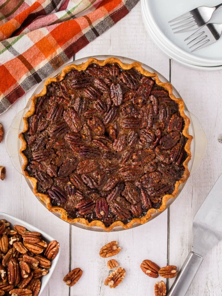 A pecan pie sits on a white wooden surface beside a knife, a bowl of pecans, stacked plates with forks, and a plaid cloth napkin.