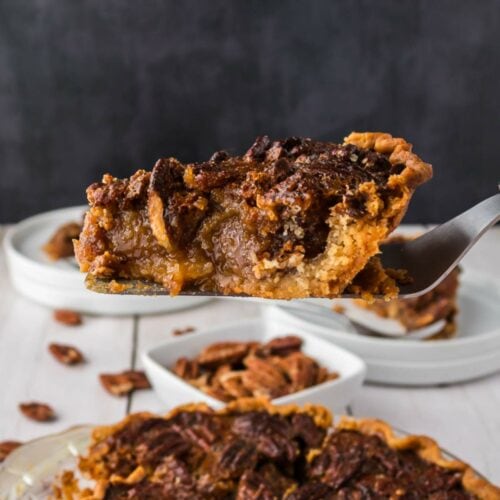 A close-up of a slice of pecan pie being lifted from a whole pie, with visible gooey filling and pecan topping.