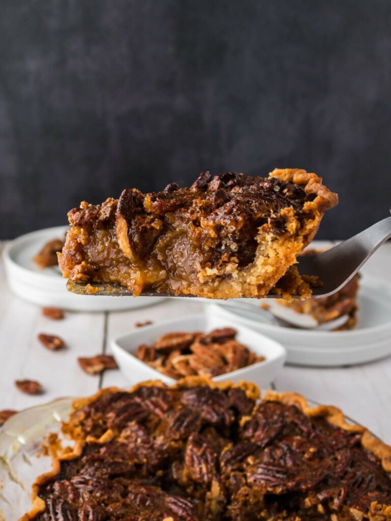 A close-up of a slice of pecan pie being lifted from a whole pie, with visible gooey filling and pecan topping.