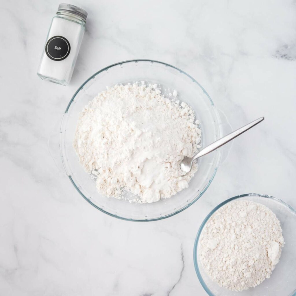 A glass bowl with flour and a spoon, sugar, and a jar of salt on a white marble surface.