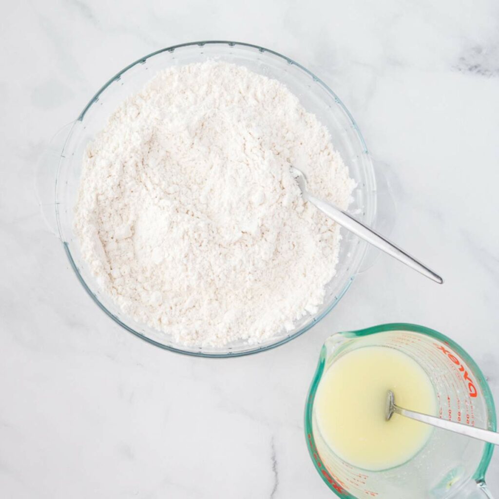 A glass bowl with flour and a spoon next to a measuring cup containing milk, both placed on a white surface.