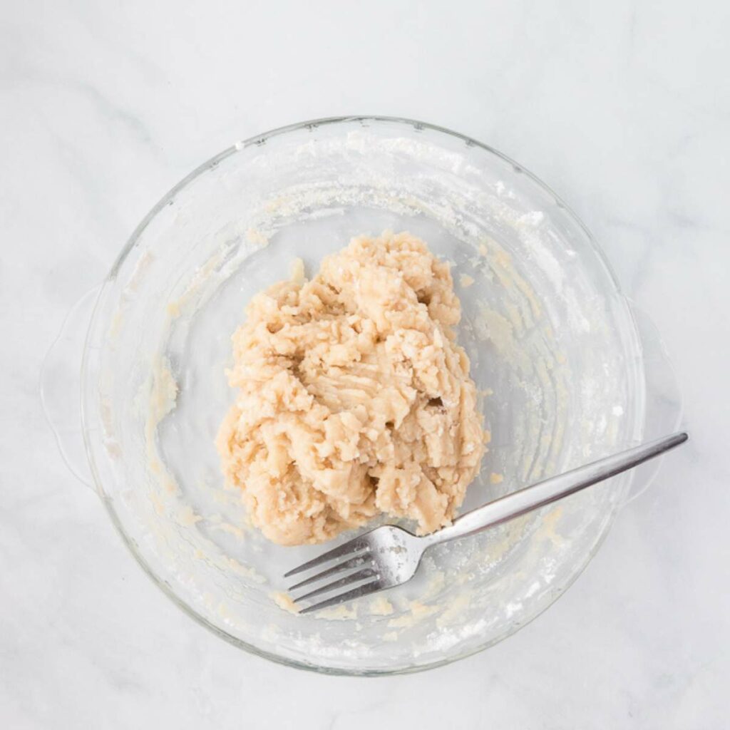 A glass bowl containing a mound of dough with a fork resting on the side, viewed from above on a white surface.