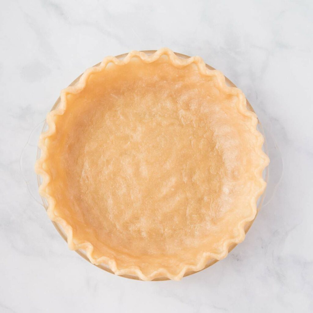 Unbaked pie crust with fluted edges in a clear glass pie dish on a light marble surface.