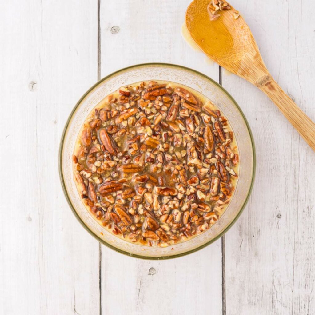A glass bowl filled with chopped pecans mixed in a liquid, next to a wooden spoon on a white wooden surface.