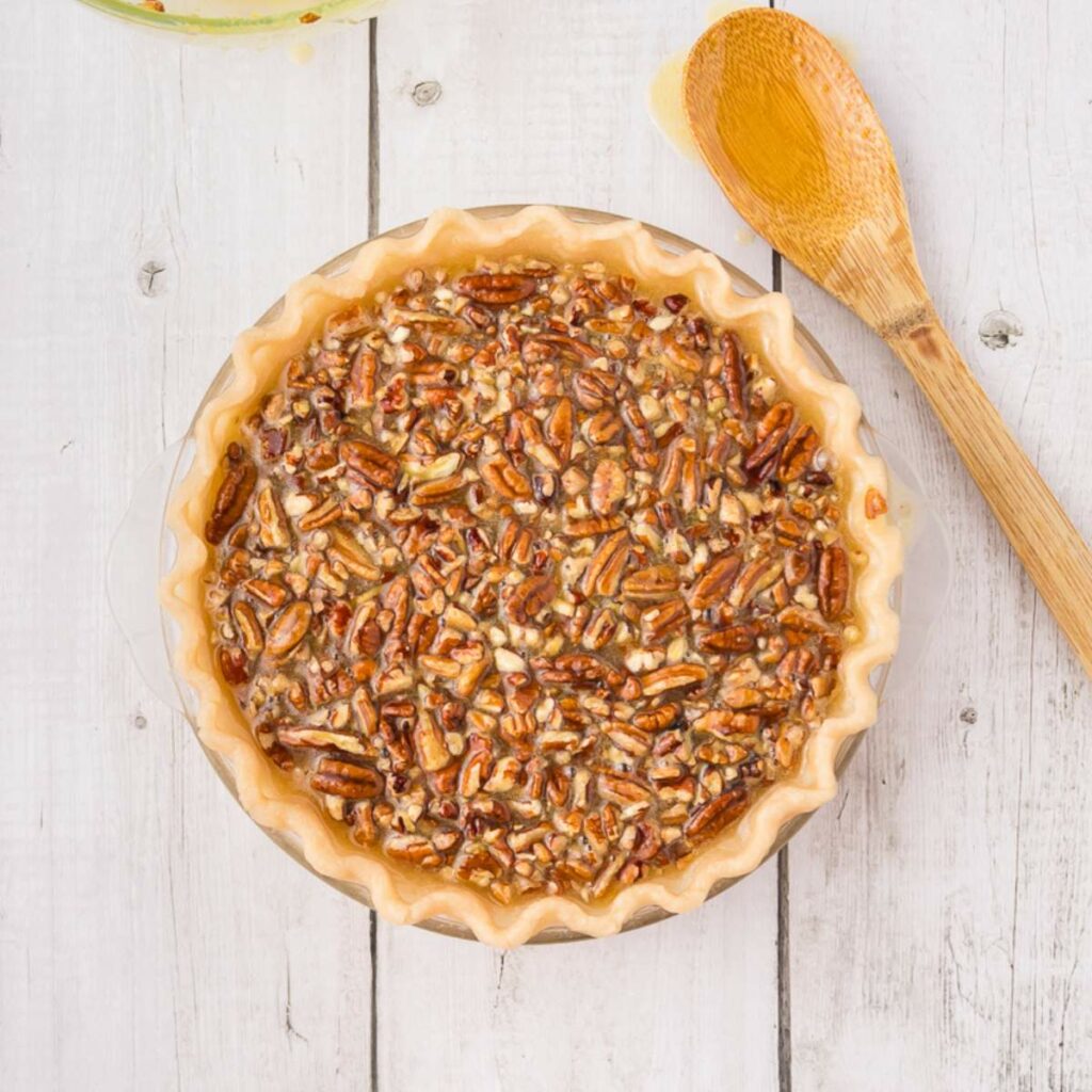 A pecan pie with an unbaked crust sits on a white wooden surface next to a wooden spoon.
