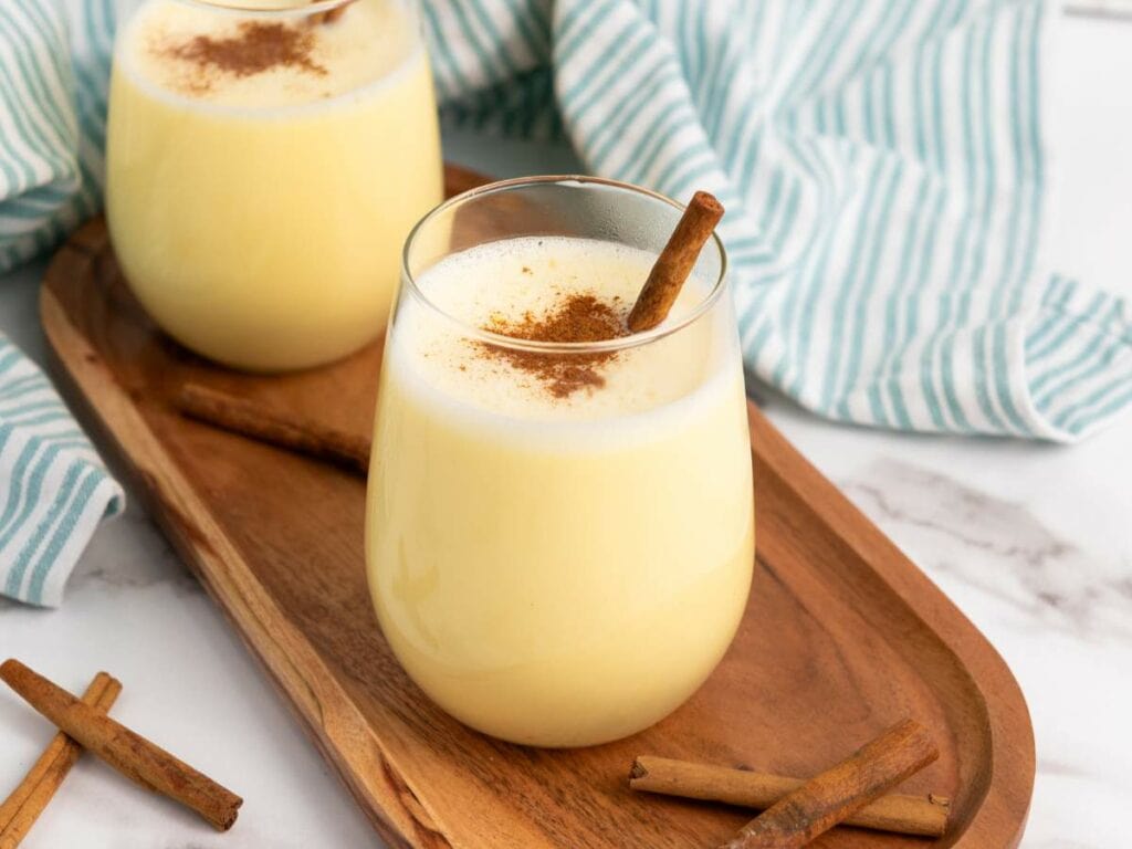 Two glasses of eggnog on a wooden tray, each topped with ground cinnamon and a cinnamon stick.