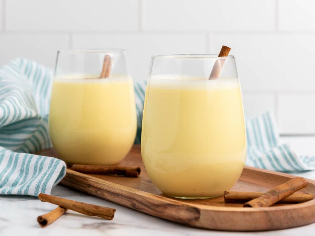 Two glasses of yellow eggnog with cinnamon sticks, placed on a wooden tray with extra cinnamon sticks and a striped cloth in the background.